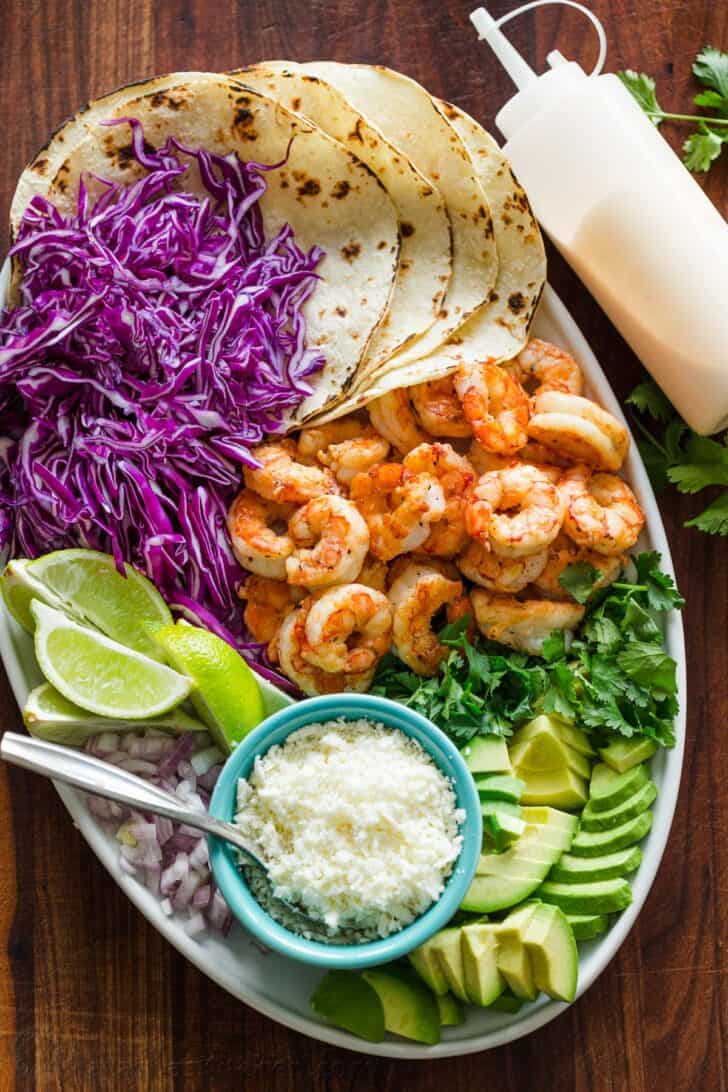 Shrimp Tacos are loaded with shrimp, cabbage, avocado, cotija, cilantro and served over gluten free corn tortillas. The shrimp taco sauce will win you over! Ingredients for serving shrimp tacos arranged on a platter with toasted tortillas