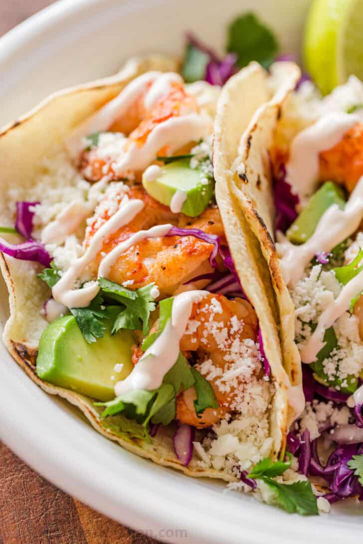Shrimp Tacos are loaded with shrimp, cabbage, avocado, cotija, cilantro and served over gluten free corn tortillas. The shrimp taco sauce will win you over! Side profile of tacos laying on platter loaded with shrimp and drizzled with sauce