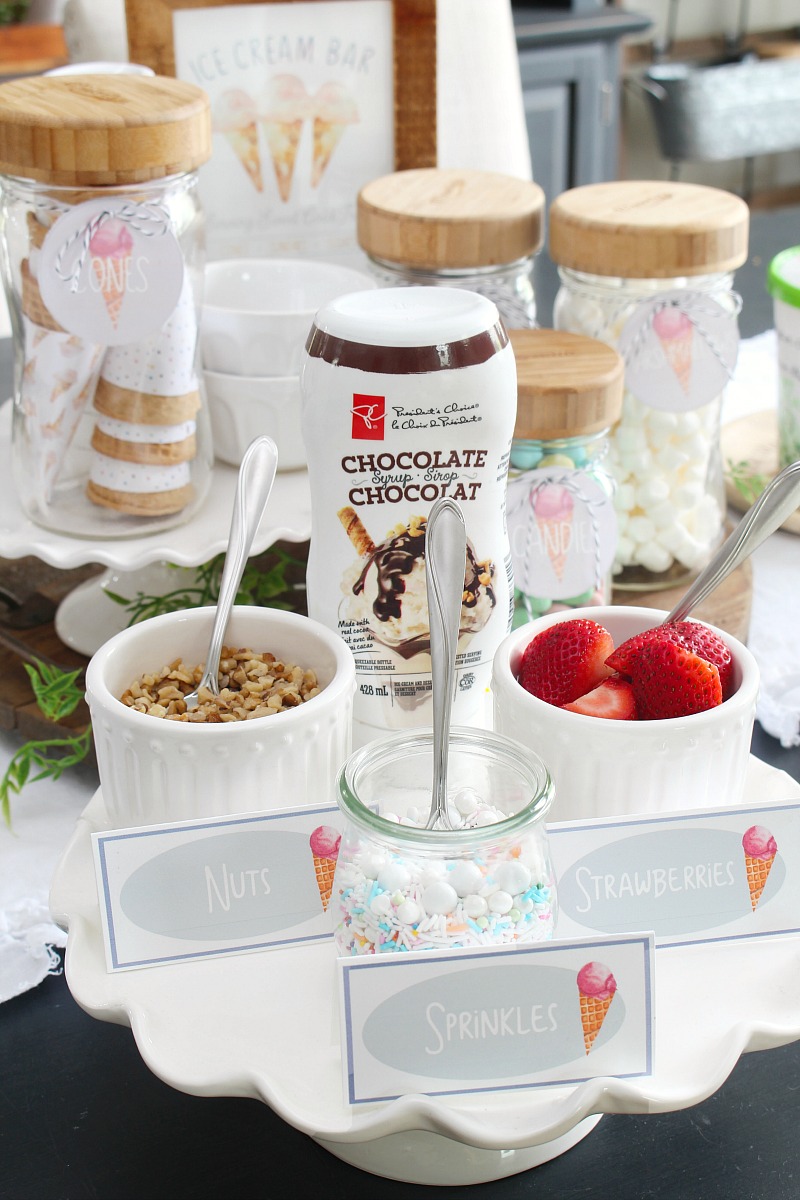 Ice Cream Sundae Bar Printables 38 Ice cream sundae bar with toppings on a cake stand.