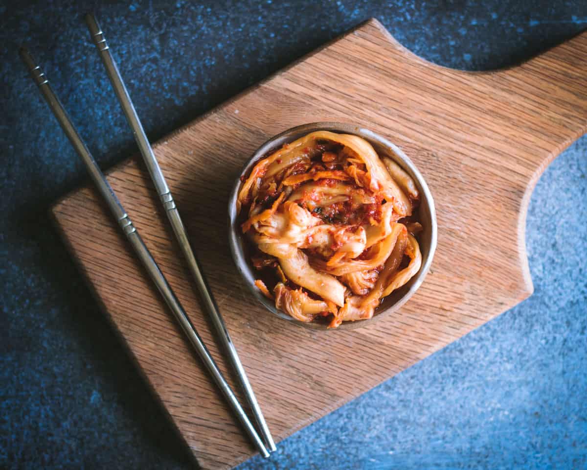 Kimchi is a spicy Korean side dish that is traditionally fermented, similar to sauerkraut. It is usually made with napa cabbage along with other vegetables like carrots, onions, ginger, and garlic. Korean chili flakes give it a spicy kick! Kimchi is surprisingly easy to make and so delicious! a small bowl of kimchi on a cutting board with chopsticks