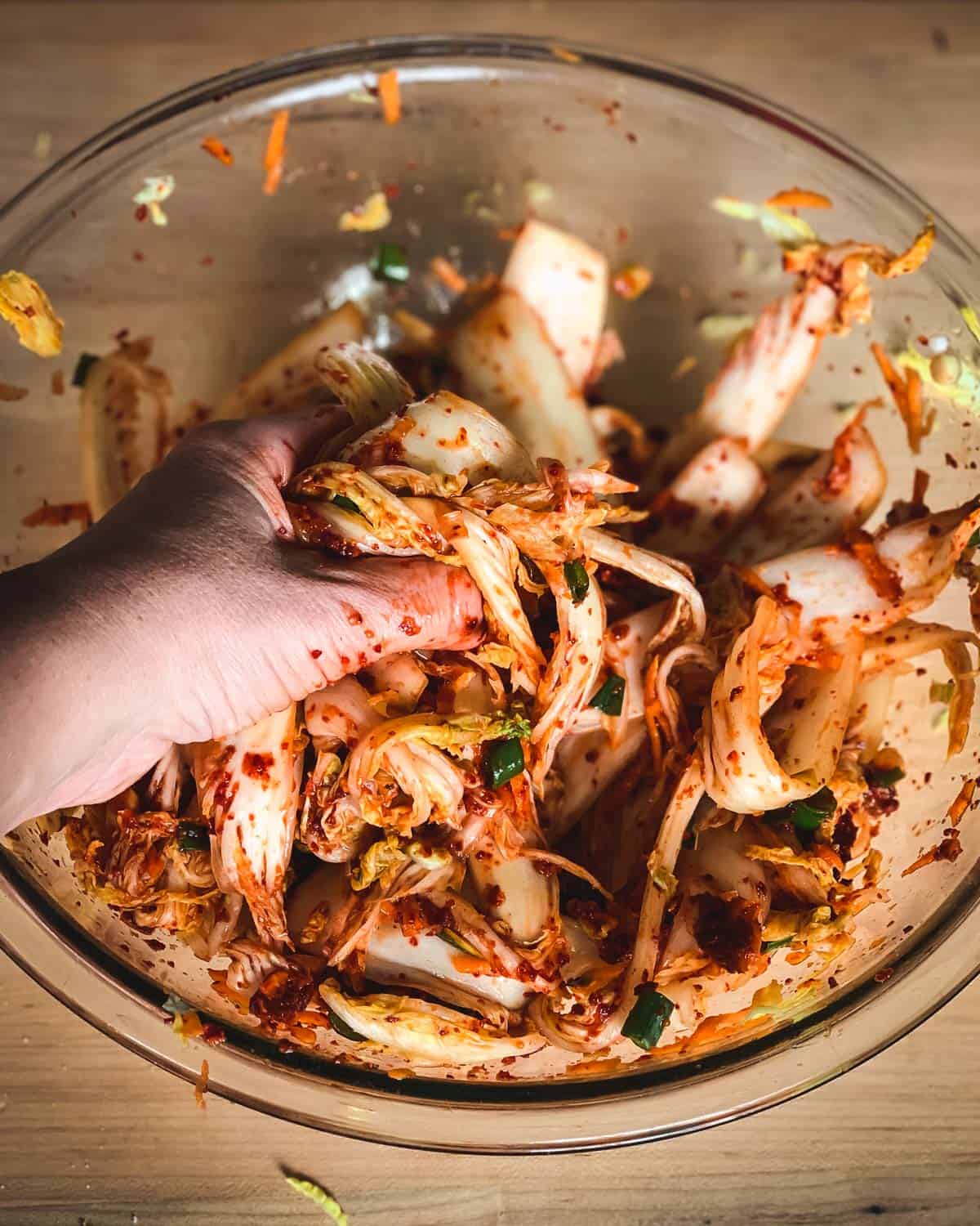 Kimchi is a spicy Korean side dish that is traditionally fermented, similar to sauerkraut. It is usually made with napa cabbage along with other vegetables like carrots, onions, ginger, and garlic. Korean chili flakes give it a spicy kick! Kimchi is surprisingly easy to make and so delicious! a hand massaging the spices into the cabbage