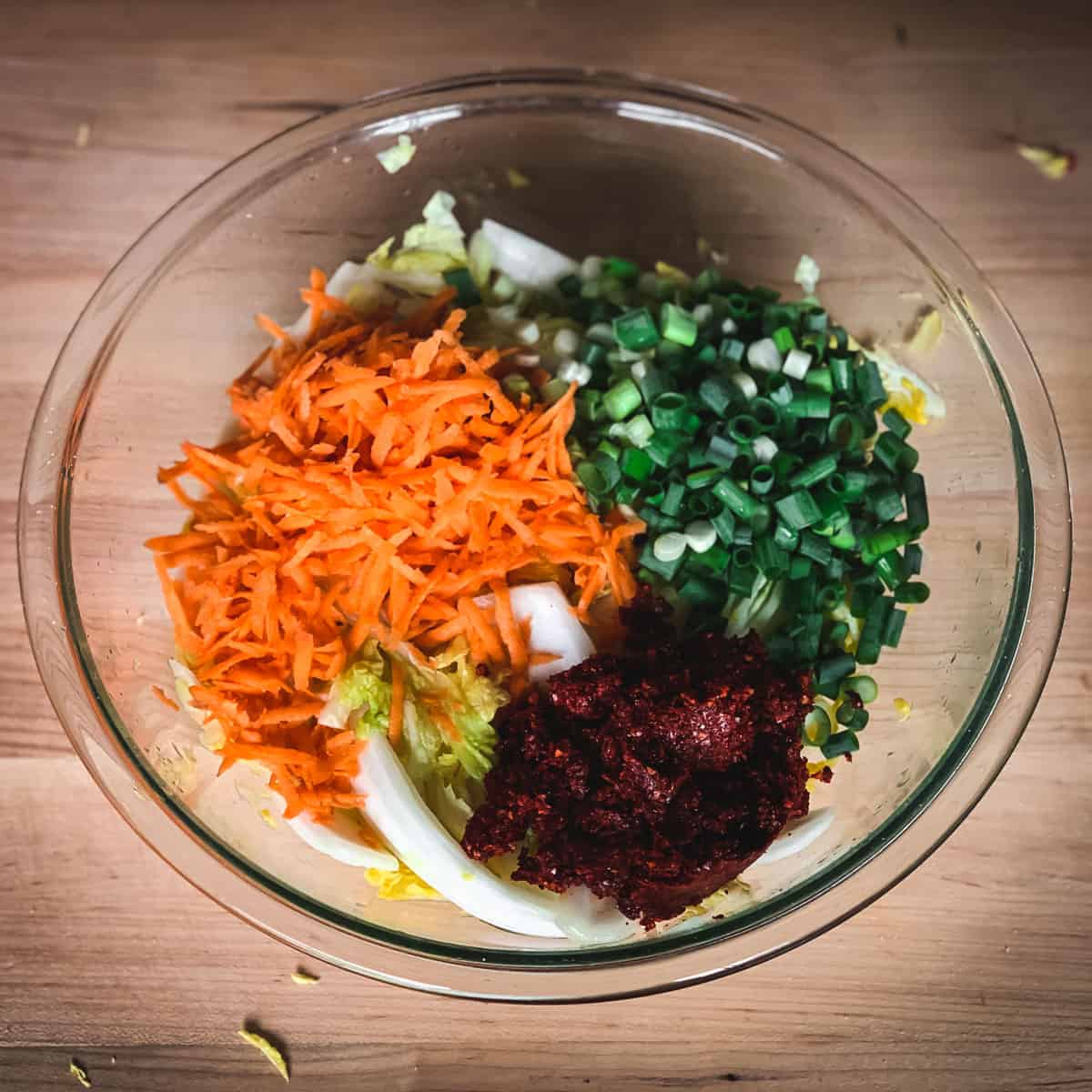 Kimchi is a spicy Korean side dish that is traditionally fermented, similar to sauerkraut. It is usually made with napa cabbage along with other vegetables like carrots, onions, ginger, and garlic. Korean chili flakes give it a spicy kick! Kimchi is surprisingly easy to make and so delicious! adding the carrots, onions, and spices to the bowl of cabbage
